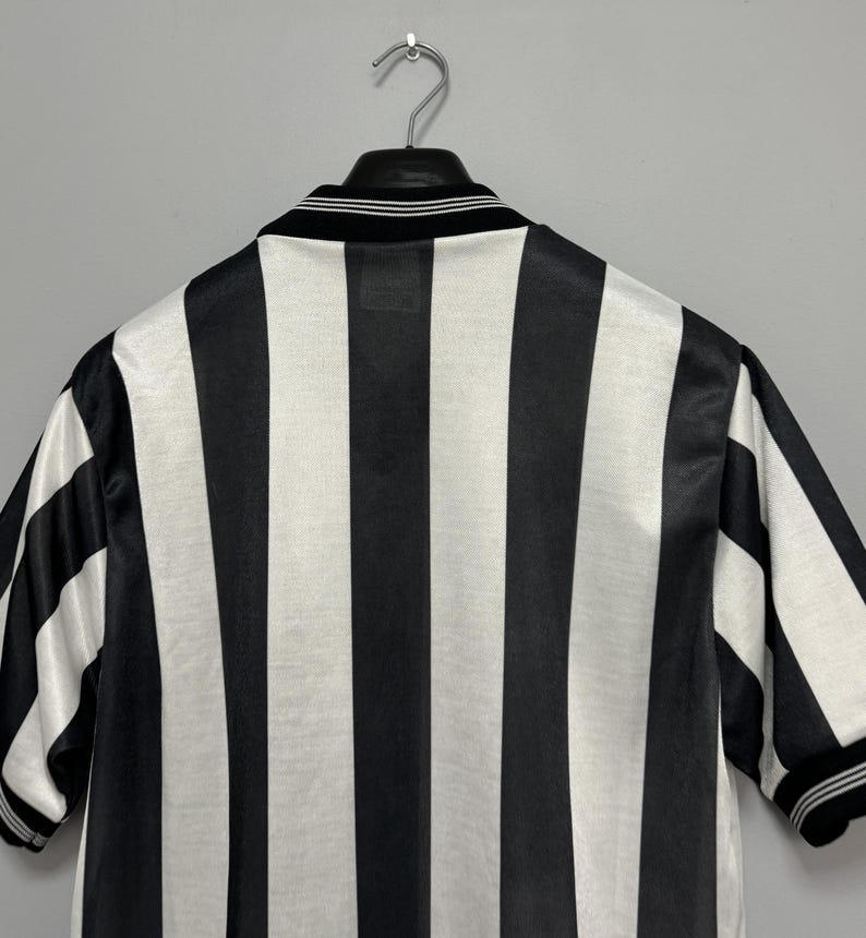 May include: Black and white striped jersey hanging on a metal hanger. The jersey has a black collar with white stripes and short sleeves. The stripes are vertical and evenly spaced.