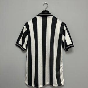 May include: Black and white striped short-sleeve jersey hanging on a metal hanger. The jersey has a collared neckline and black trim on the sleeves and collar. The stripes are vertical and evenly spaced.