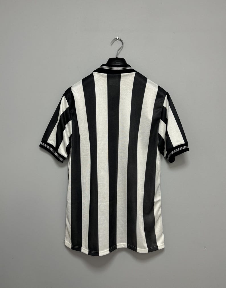 May include: Black and white striped short-sleeve jersey hanging on a metal hanger. The jersey has a collared neckline and black trim on the sleeves and collar. The stripes are vertical and evenly spaced.