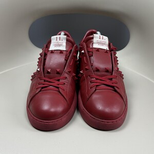 May include: A pair of red leather low-top trainers with a studded design. The trainers have a white label on the side that reads "11. ROCKSTUD UNTITLED  ROMAN", and the laces are red.