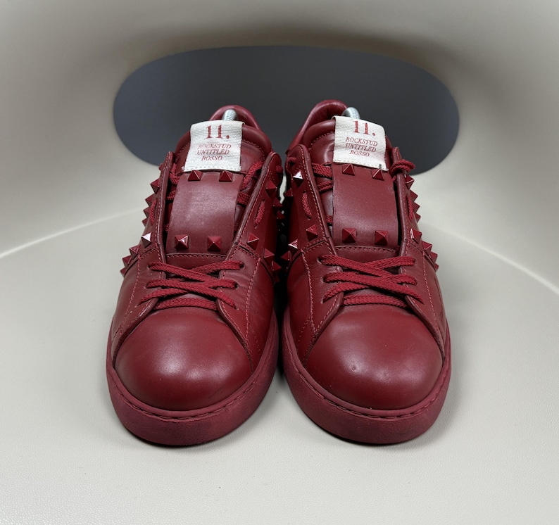 May include: A pair of red leather low-top trainers with a studded design. The trainers have a white label on the side that reads "11. ROCKSTUD UNTITLED  ROMAN", and the laces are red.