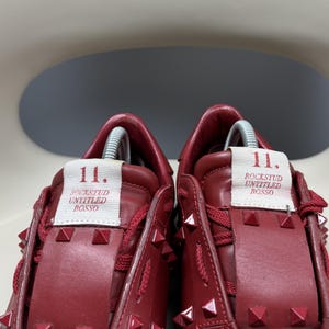 May include: A pair of red leather trainers with silver studs. The trainers have a white label on the side that says "11. Rockstud Untitled Rosso."