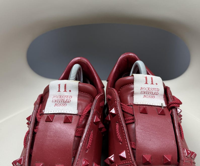 May include: A pair of red leather trainers with silver studs. The trainers have a white label on the side that says "11. Rockstud Untitled Rosso."