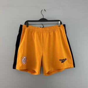 May include: Gold athletic shorts with black side panels and an elastic waistband. The front features a circular team crest on the right leg and a black Reebok logo on the left leg. The shorts are displayed on a black plastic clothes hanger.