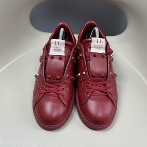 May include: A pair of red leather trainers with a studded design. The trainers have a white label on the side that says "Rockstud Untitled Rosso 11."