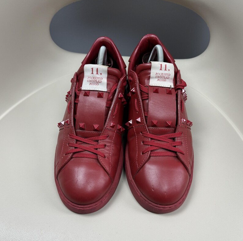 May include: A pair of red leather trainers with a studded design. The trainers have a white label on the side that says "Rockstud Untitled Rosso 11."