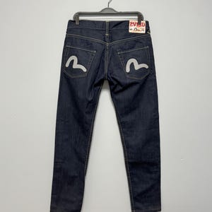 Puede incluir: Vaqueros de mezclilla azul índigo oscuro con un logotipo de gaviota pintado en blanco en cada bolsillo trasero y una etiqueta de marca rectangular blanca en la cintura trasera con el texto rojo Evisu y detalles negros, en una percha negra.