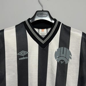 May include: Black and white striped soccer jersey with a V-neck collar and the Umbro logo. The jersey has a circular emblem on the right side and a label that reads "107cm 42 inch".