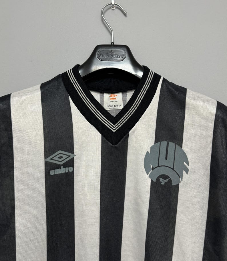 May include: Black and white striped soccer jersey with a V-neck collar and the Umbro logo. The jersey has a circular emblem on the right side and a label that reads "107cm 42 inch".