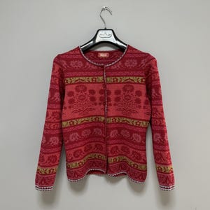 Peut inclure: Cardigan rouge à motifs floraux et manches longues. Le pull boutonné a un col rond et est suspendu à un cintre noir. La bordure est blanche.