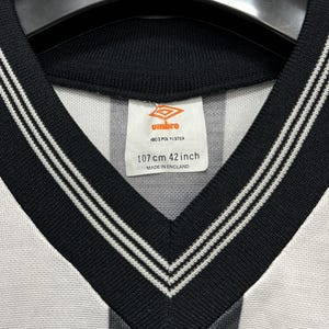 May include: Close-up of a vintage Umbro soccer jersey. The jersey has a black and white striped design with a black V-neck collar and white trim. The tag reads "107 cm 42 inch" and "Made in England."