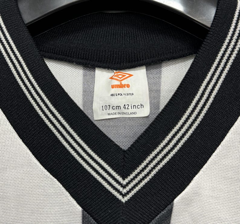May include: Close-up of a vintage Umbro soccer jersey. The jersey has a black and white striped design with a black V-neck collar and white trim. The tag reads "107 cm 42 inch" and "Made in England."