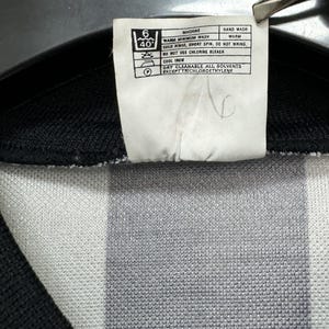 May include: Close-up of a white tag with care instructions for a garment. The tag includes washing symbols and text indicating machine wash, cold rinse, and cool iron. The garment has a black and white striped pattern.