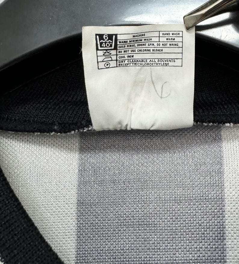 May include: Close-up of a white tag with care instructions for a garment. The tag includes washing symbols and text indicating machine wash, cold rinse, and cool iron. The garment has a black and white striped pattern.