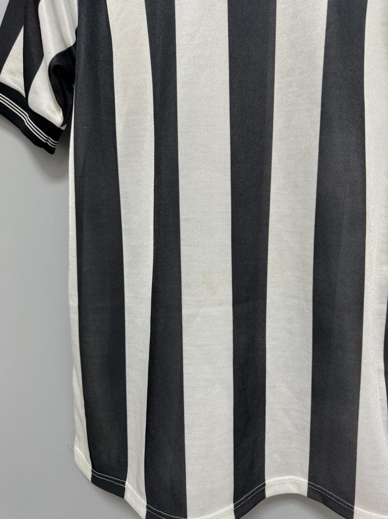 May include: Close-up of a black and white striped shirt. The shirt has vertical stripes and a white collar with black trim. The fabric appears to be a breathable material, suitable for athletic wear or casual use.