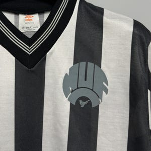 May include: A black and white striped jersey with a V-neck collar. The jersey features a gray emblem on the front and the text "107cm 42 inch" near the collar. The jersey is made of a lightweight material.