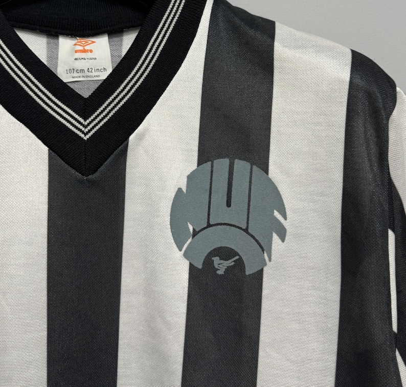 May include: A black and white striped jersey with a V-neck collar. The jersey features a gray emblem on the front and the text "107cm 42 inch" near the collar. The jersey is made of a lightweight material.