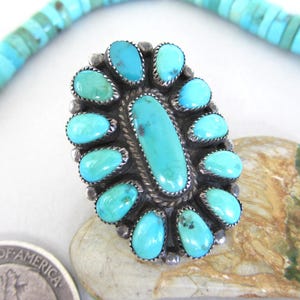 May include: Oval-shaped turquoise ring with a silver setting. The ring features a large, central turquoise stone surrounded by smaller turquoise stones. A turquoise bead necklace is visible in the background.