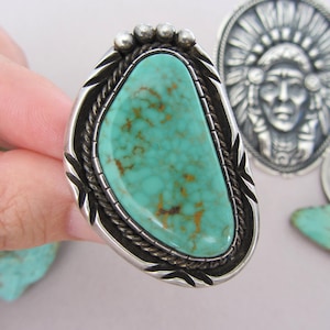 May include: A large, silver-toned ring featuring a vibrant, turquoise stone with brown veining. The ring has a decorative border with a rope-like design and leaf accents. Additional silver jewelry pieces are visible in the background.