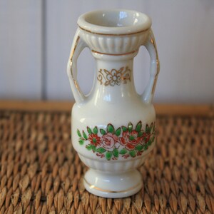 Op de afbeelding: Een kleine witte porseleinen vaas met gouden accenten en bloemmotieven. De vaas heeft twee handvatten en een ronde basis.