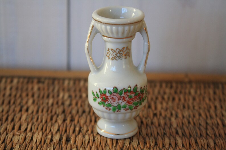 Op de afbeelding: Een kleine witte porseleinen vaas met gouden accenten en bloemmotieven. De vaas heeft twee handvatten en een ronde basis.