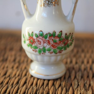 Op de afbeelding: Een kleine witte porseleinen vaas met een bloemenmotief. De vaas heeft twee handvatten en een gouden rand rond de bovenkant.