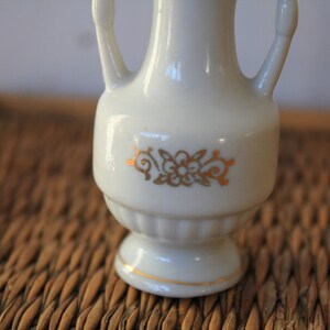 Op de afbeelding: Een witte keramische vaas met gouden bloemmotieven en twee handvatten. De vaas staat op een geweven rieten oppervlak.