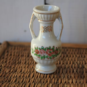Op de afbeelding: Een kleine witte porseleinen vaas met gouden accenten en een bloemenontwerp. De vaas heeft twee handvatten en een ronde basis.