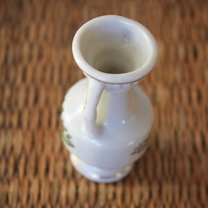 Op de afbeelding: Een kleine witte keramische vaas met een gouden rand en bloemmotieven. De vaas heeft een handvat en een rond lichaam.