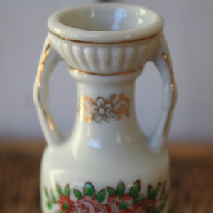 Op de afbeelding: Een kleine witte porseleinen vaas met gouden accenten en bloemmotieven. De vaas heeft twee handvatten en een brede opening.