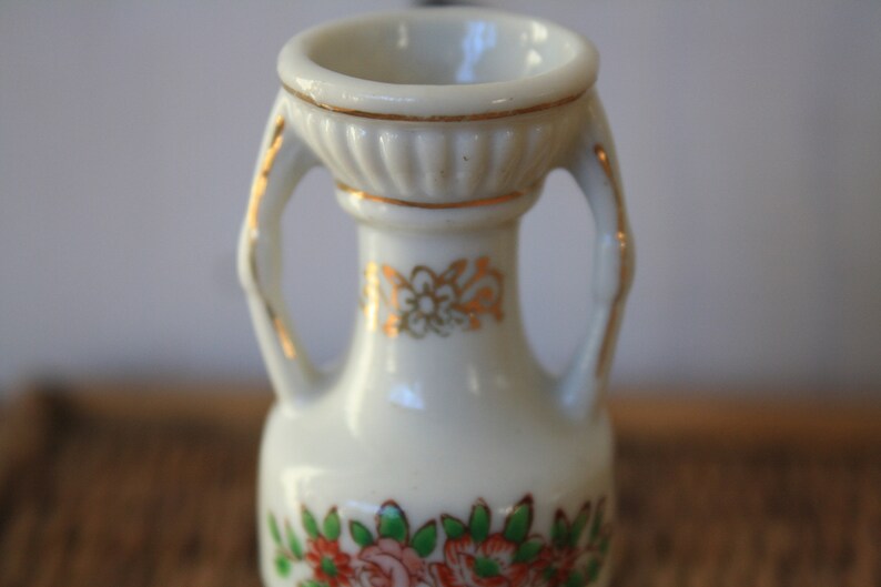 Op de afbeelding: Een kleine witte porseleinen vaas met gouden accenten en bloemmotieven. De vaas heeft twee handvatten en een brede opening.