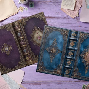 May include: Two antique-style, open books with ornate gold detailing and floral designs. One book has a deep purple cover, the other a teal blue cover. The books are on a light purple wooden surface, with vintage paper scraps scattered around.