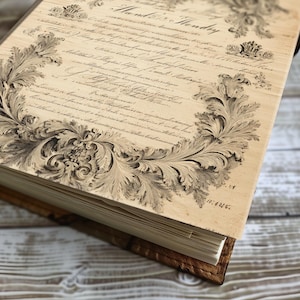 May include: A vintage-style journal with aged paper pages and a decorative, ornate border. The cover features handwritten script and a detailed floral design in shades of brown and beige. The journal is bound in a brown leather-like material.