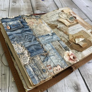 May include: A handmade journal with a denim and lace cover. The cover is decorated with various fabric scraps, including floral patterns and a delicate lace trim. The journal is open to reveal pages filled with handwritten notes and ephemera.