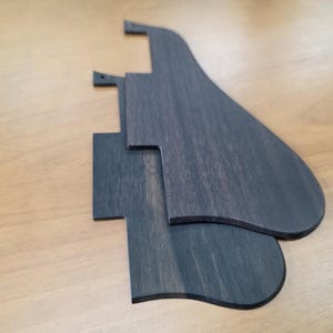 Puede incluir: Tres protectores de guitarra de madera gris oscuro apilados sobre una superficie marrón claro. Los protectores tienen un acabado liso y están cortados en diferentes formas, incluyendo una lágrima y una forma rectangular.