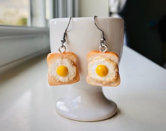 May include: A pair of silver dangle earrings with a fried egg on toast charm. The toast is light brown and the egg yolk is yellow.