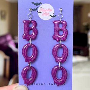 Peut inclure: Boucles d'oreilles pendantes violettes scintillantes formant le mot "BOO". Ces boucles d'oreilles faites à la main sur le thème d'Halloween sont reliées par des anneaux argentés.
