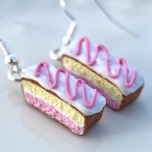 May include: A pair of cake slice earrings. Each earring is a miniature replica of a cake slice, with a brown base, pink and yellow layers, white icing, and a pink zigzag pattern. They hang from silver hooks.