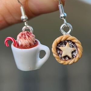 May include: A pair of novelty earrings. One earring is a white mug of hot chocolate with whipped cream, cocoa powder, and a candy cane. The other earring is a miniature pie with a star cutout and a braided crust.