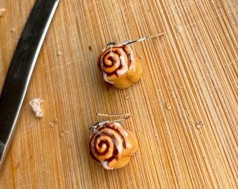 Puede incluir: Un par de pendientes de rollo de canela con ganchos plateados. Los pendientes están hechos de arcilla polimérica y tienen forma de rollo de canela con glaseado blanco y remolinos marrones.