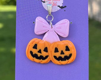 May include: A silver necklace with a pendant featuring two orange jack-o'-lanterns with black carved faces, a pink bow, and a small blue gem. The pendant is on a purple background with bat silhouettes.