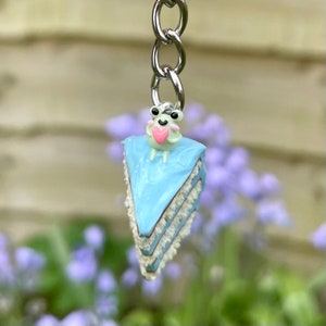 Puede incluir: Un llavero caprichoso suspendido de una cadena plateada. Presenta una porción de pastel azul, coronada por una pequeña rana que sostiene un corazón rosa. El pastel tiene glaseado blanco y un glaseado azul claro. El fondo está borroso con tonos verdes y morados.
