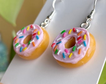 May include: A pair of donut earrings with pink icing and colourful sprinkles. The earrings are made of polymer clay and have silver hooks.