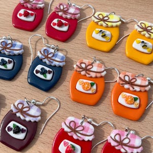 Op de afbeelding: Een collectie miniatuur oorbellen in de vorm van potjes met diverse fruit met diverse ontwerpen zoals aardbei, bosbes, sinaasappel en braam. Elke oorbel heeft een deksel met ruitjespatroon, een bruin strikje en zilverkleurige metalen haken.