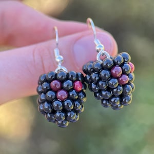 May include: A pair of handmade earrings shaped like blackberries. Each earring features a cluster of small, glossy black beads with a few red beads interspersed. The earrings have silver-colored fish hook ear wires.