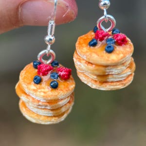 May include: A pair of drop earrings featuring miniature stacks of golden-brown pancakes topped with glossy blue blueberries, bright red raspberries, and a translucent amber syrup glaze, all suspended from silver-toned metal earring hooks.