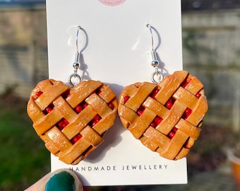 May include: Heart-shaped earrings resembling miniature cherry pies. The earrings feature a lattice crust design with visible red cherry filling. The earrings are handmade jewelry with silver-colored hooks. The text "Auntie Abi Jewellery" is visible on the card.