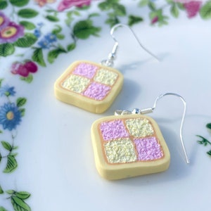 May include: A pair of silver dangle earrings featuring a square-shaped charm with a yellow and pink checkerboard design. The charm resembles a miniature cake.