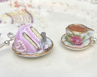 Puede incluir: Un par de pendientes con tazas de té y rebanadas de pastel de porcelana en miniatura sobre platos pequeños. Las tazas de té están decoradas con rosas rosas y ribetes dorados. Las rebanadas de pastel son rosas con glaseado blanco y ribetes dorados.