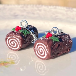 May include: A pair of earrings shaped like Yule log cake with chocolate frosting, white filling, and red holly berries.